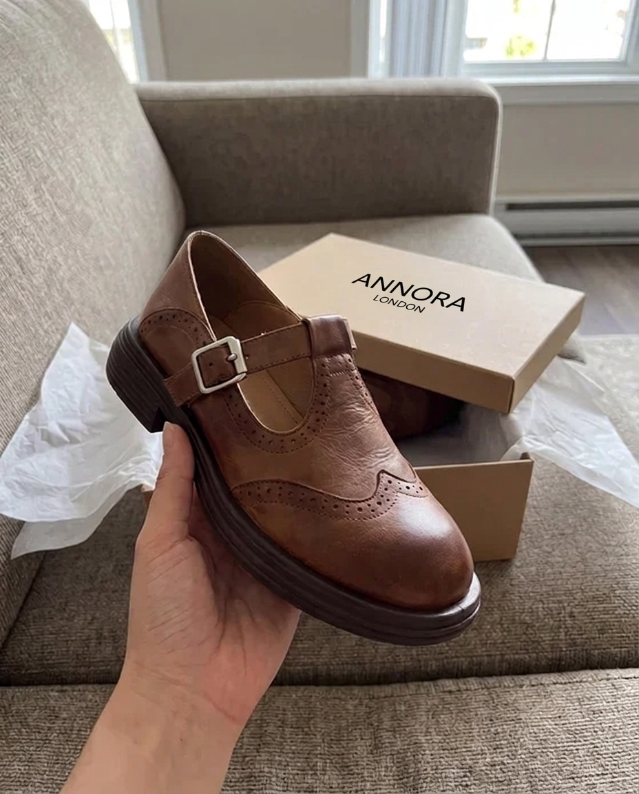 Brown brogue T-bar shoe held in hand beside its ANNORA LONDON box