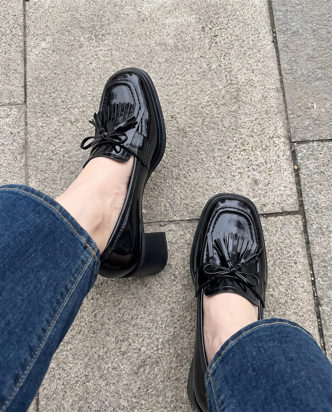 Black patent fringed loafers worn with jeans on a pavement